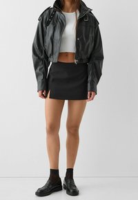 Veste cropped en simili cuir noire, top court à rayures blanches, mini-jupe noire avec fente latérale et mocassins noirs avec des chaussettes transparentes.