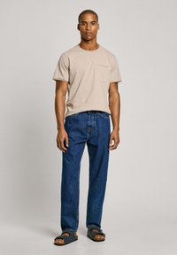 Camiseta de algodón beige con bolsillo, jeans de pierna recta azul oscuro y sandalias negras. El modelo está de pie contra un fondo gris claro.