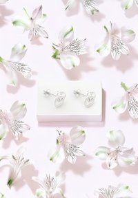 Silberne herzförmige Steckerohrringe auf einer weißen Box präsentiert, umgeben von zarten rosa Blumen mit grünen Akzenten auf einem sanften rosafarbenen Hintergrund.