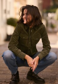 Camicia in velluto a coste verde oliva con due tasche sul petto, bottoni metallici e maniche lunghe, abbinata a jeans blu e stivali neri chunky.