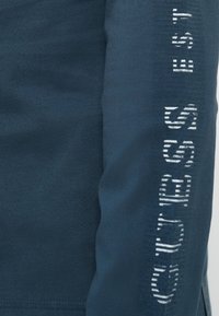 Niebieska bluzka z długim rękawem wykonana z miękkiego materiału, z wyraźnym białym logo "GUESS" w teksturowanym wzorze, umieszczonym pionowo wzdłuż prawego rękawa.
