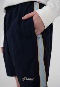 Gros plan d'une main dans la poche d'un short bleu marine avec des bandes latérales bleu clair et jaune, et le mot « Prohibited » brodé près de l'ourlet.