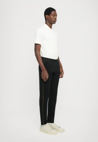 Chemise polo blanche à manches courtes, pantalon noir avec des rayures jaunes sur le côté, et baskets crème. Design simple avec une silhouette ajustée.