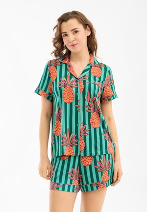 Joven mujer vestida con pijama de rayas verdes de manga corta con estampados de piñas naranjas brillantes, de pie con expresión neutral.