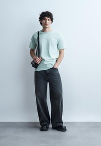 Mladý muž stojí opřený o hladkou zeď, má na sobě světle zelené tričko, volné černé džíny, černé boty a nese malou černou kabelku přes rameno.