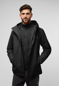 Zwarte waterdichte jas met een capuchon, gelaagd over een zwart fleece innerlijk. Voorzien van een ritssluiting aan de voorkant en twee zijzakken.