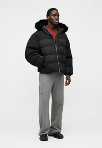 Μαύρο μπουφάν puffer με κουκούλα από ψεύτικη γούνα, κλείσιμο με φερμουάρ και σχέδιο καπιτονέ. Συνδυασμένο με γκρι φόρμες και μαύρα αθλητικά παπούτσια.