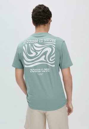 Mintgroene katoenen t-shirt met een witte golvende grafische tekening en tekst op de achterkant. Korte mouwen en een ronde halslijn.