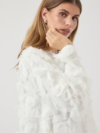 Blouse blanche avec une finition texturée présentant des détails frangés. Le tissu est doux, avec de longues et fines bandes créant un effet superposé.