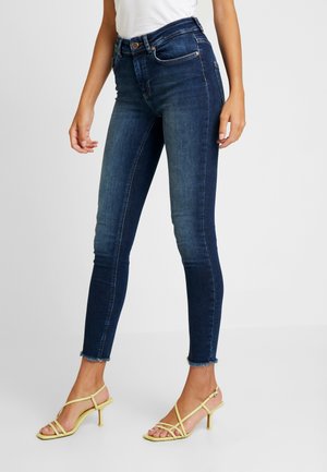 Jeans ajustados azul oscuro con cintura alta, que presentan una textura desgastada y dobladillo deshilachado. Combinados con tacones amarillos con tiras.