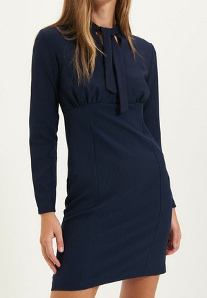 Vrouw draagt een marineblauwe, langarmige jurk tot op de knie met een gestrikte kraagdetail tegen een effen achtergrond.