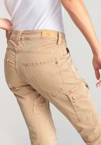 Pantalones cargo beige con un acabado texturizado, presentan múltiples bolsillos, una etiqueta en la cinturilla y detalles en tono dorado.