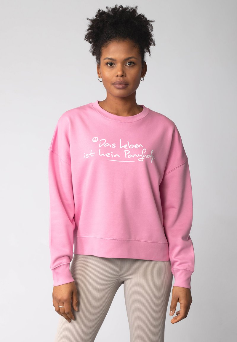 Der pinkfarbene Sweatshirt hat lange Ärmel, einen weiten Ausschnitt und weißen Text in einer lässigen Schriftart. Kombiniert mit hellen, gerippten Leggings.