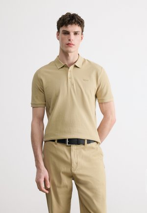 Młody mężczyzna w beżowej koszulce polo i dopasowanych spodniach z czarnym paskiem, stojący na jednolitym białym tle.