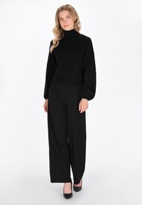 Zwarte coltrui met ballonmouwen, gecombineerd met zwarte broek met wijde pijpen. De outfit heeft een textuurstof en een strakke uitstraling.