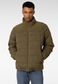 Mann iført olivengrønn zip-up pufferjakke med hendene i lommene, stående mot en enkel lys bakgrunn.