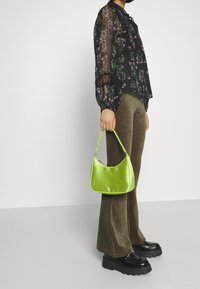 Borsa in raso verde con forma curvy e manici corti, abbinata a un top nero in chiffon floreale e pantaloni in corduroy olive. Scarpe con plateau nere.