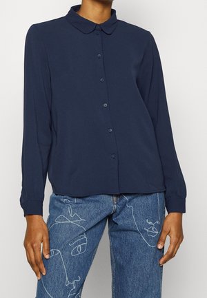 Marineblaue Langarmbluse mit Kragen, ausgestattet mit einer durchgehenden Knopfleiste und einer weichen Textur. Kombiniert mit hellblauen Jeans mit einem Gesichtskonturenmuster.