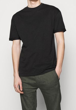 Personne portant un T-shirt noir uni à manches courtes et un pantalon vert foncé, une main dans la poche, debout devant un fond neutre.