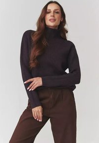 Kobieta z długimi falującymi włosami, ubrana w ciemny sweter z prążkowanym golfem i brązowe spodnie, pozująca z założonymi na biodrach rękami na tle jednolitego tła.