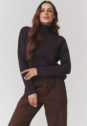 Kobieta z długimi falującymi włosami, ubrana w ciemny sweter z prążkowanym golfem i brązowe spodnie, pozująca z założonymi na biodrach rękami na tle jednolitego tła.