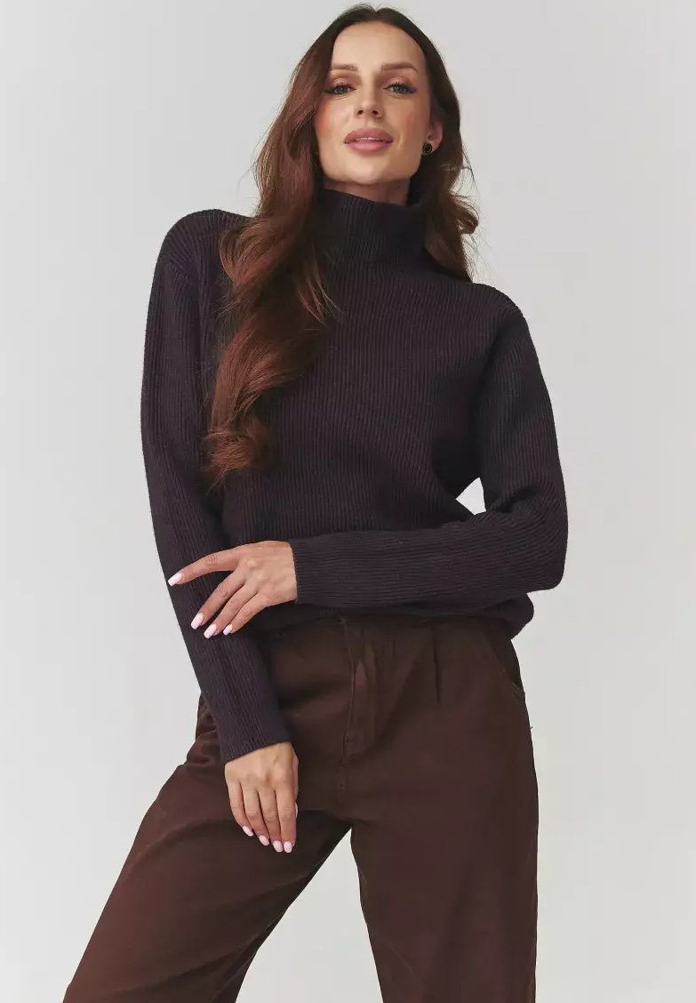 Kobieta z długimi falującymi włosami, ubrana w ciemny sweter z prążkowanym golfem i brązowe spodnie, pozująca z założonymi na biodrach rękami na tle jednolitego tła.
