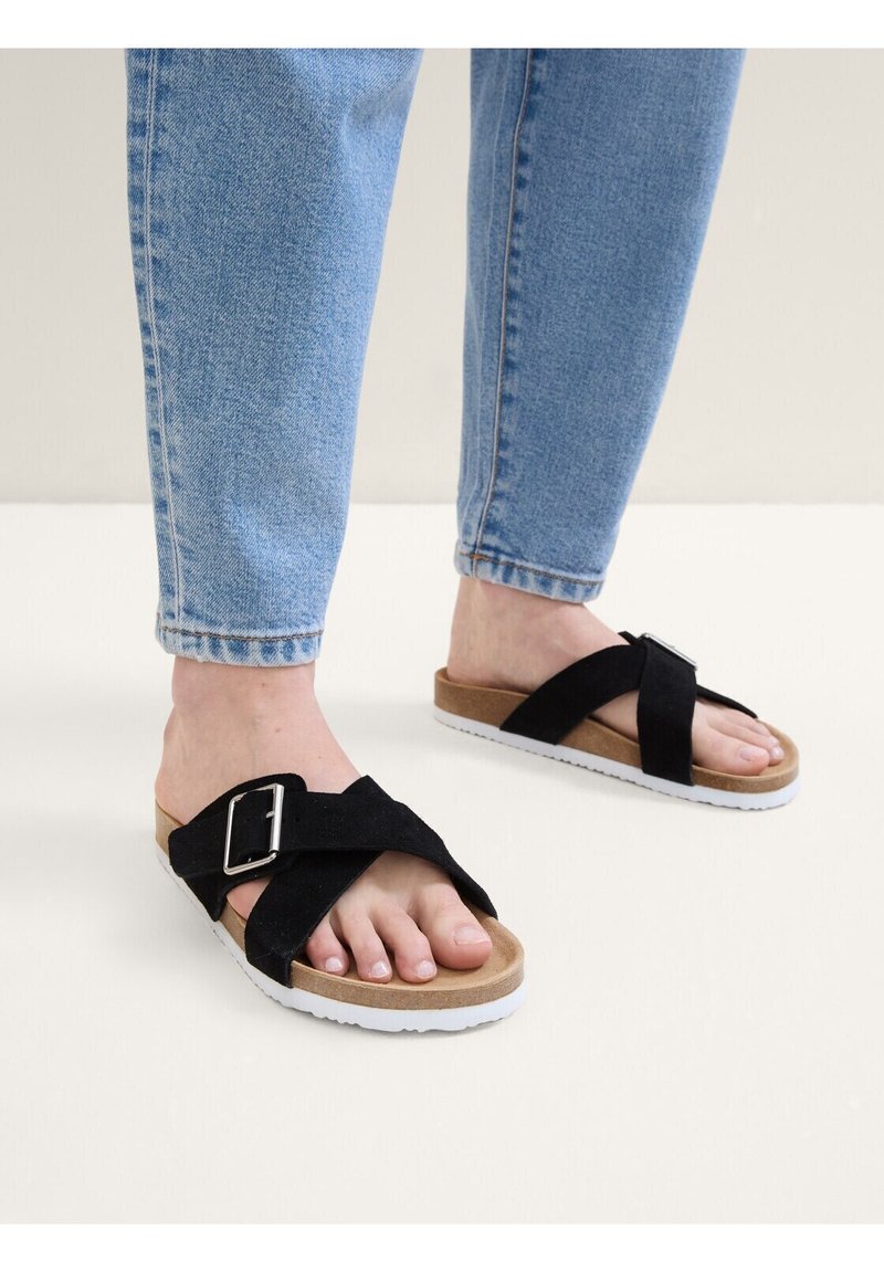 Svarta mocka slide-sandaler med en korsremdesign och metallspänne, placerade på en korkfotbädd och vit gummisula.