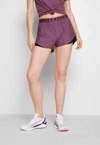 Pantalones cortos deportivos morados con detalles en negro y una cinturilla con la marca, combinados con zapatillas blancas con detalles en púrpura y rosa.