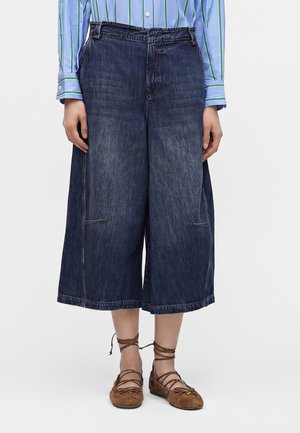 Persona che indossa pantaloni cropped in denim scuro a gamba larga, una camicia a righe blu e ballerine marroni con lacci.