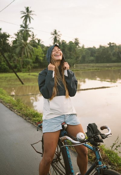 Lättviktsjacka med huva i mörkgrått och vitt, kombinerad med denimshorts. Cykeln är blå med en silverfärgad klocka. Lummig grönska och vatten i bakgrunden.