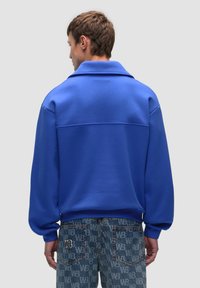Felpa blu con vestibilità rilassata e ampio colletto, realizzata in tessuto morbido. Abbinata a jeans in denim blu con motivo e design del logo.