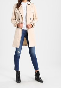 Kobieta w beżowym płaszczu, białej koszuli, podartych niebieskich dżinsach i czarnych botkach na kostkę stoi, dostosowując przód płaszcza.