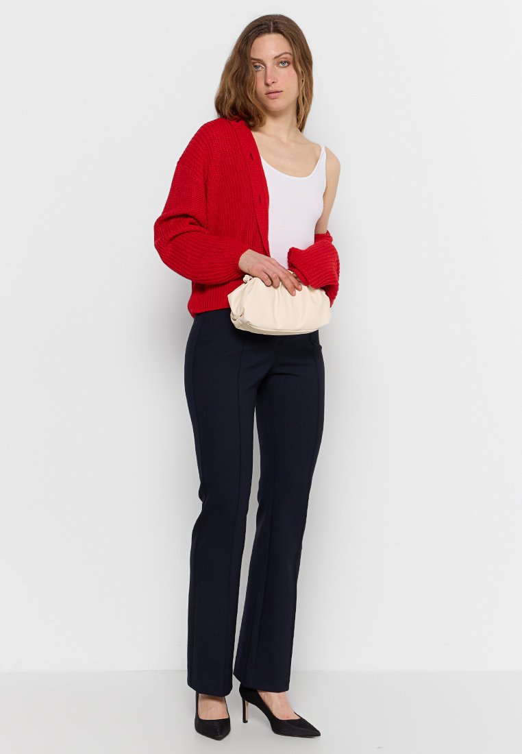Cardigan en tricot rouge, débardeur blanc, pantalon bleu marine ajusté et talons noirs. Tenant un petit sac à main crème. Tenue simple et élégante.