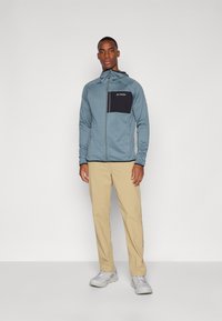 Giacca leggera blu con zip e tasca nera sul petto, abbinata a pantaloni beige e scarpe grigie. Presenta un cappuccio e tasche con zip.