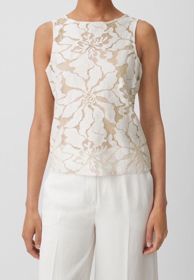 Blomstermönstrad spetsblus i beige och kräm, ärmlös design, rund halsringning, semi-genomskinlig textur, tillsammans med vita vida byxor.