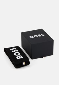 Juodas stačiakampis dėžutė su baltu "BOSS" logotipu; yra sulankstyta juoda rankšluostį su tuo pačiu logotipu ir mažu auksiniu akcento ženkleliu.
