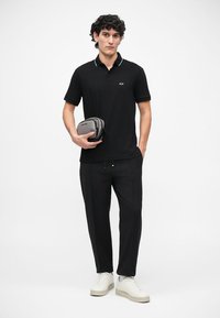Crna polo majica s bijelim detaljima na ovratniku, uparen s crnim hlačama i bijelim tenisicama. Model drži malu sivu torbu. Jednostavan dizajn.