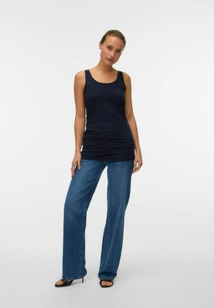 Woman standing against white background wearing sleeveless navy top, blue wide-leg jeans, and black open-toe heeled sandals.