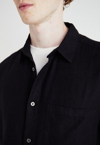Camisa negra de botones con cuello, bolsillo frontal y botones plateados. Exhibida en un modelo con una camiseta blanca debajo.