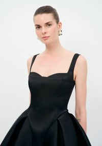 Robe en satin noir avec un corsage ajusté, des bretelles larges et une jupe plissée évasée. Texture lisse avec un décolleté en cœur.