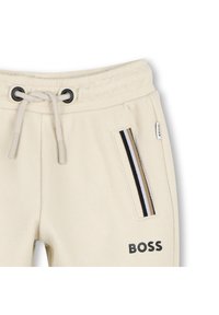 Pantalon de survêtement beige texturé avec taille à cordon, poche latérale ornée de rayures noires, blanches et fauves, et logo "BOSS" imprimé près de la poche.