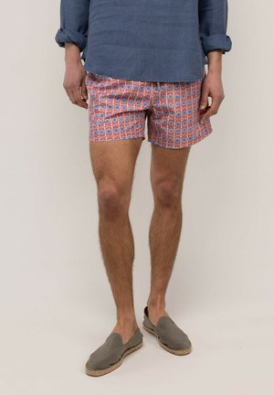 Hombre con pantalones cortos rosas estampados, camisa azul con mangas remangadas y zapatos grises sin cordones, de pie sobre un fondo liso.