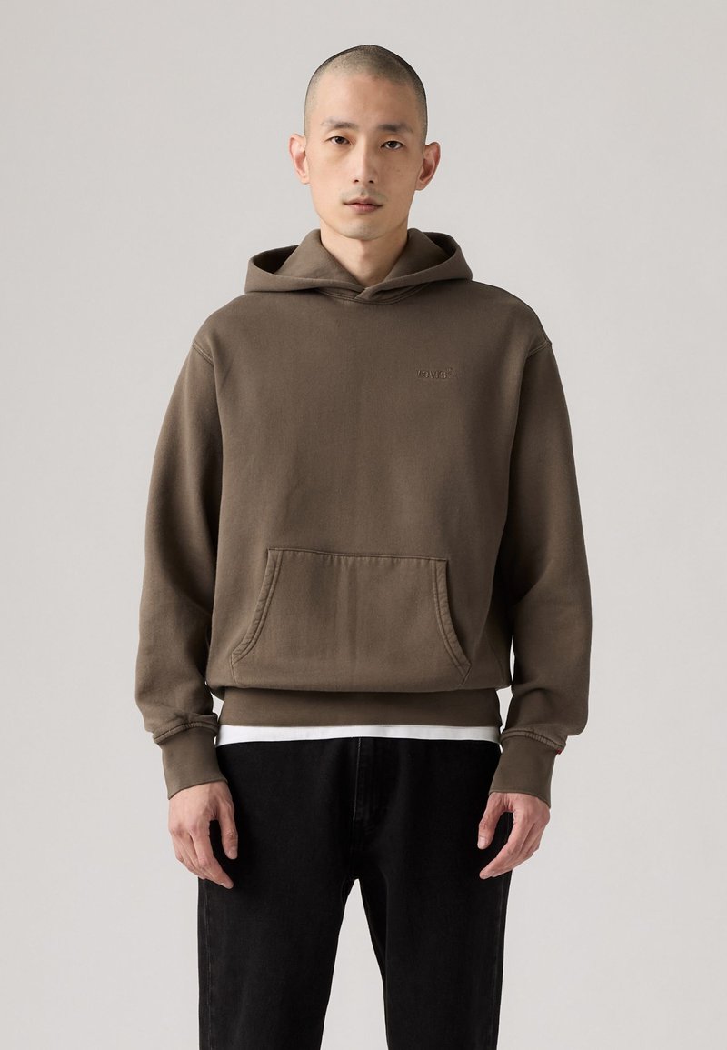 Brauner Baumwollhoodie mit einer Kängurutasche, gerippten Bündchen und einem kleinen Logo auf der Brust. Getragen über einem weißen Shirt und kombiniert mit schwarzen Hosen.