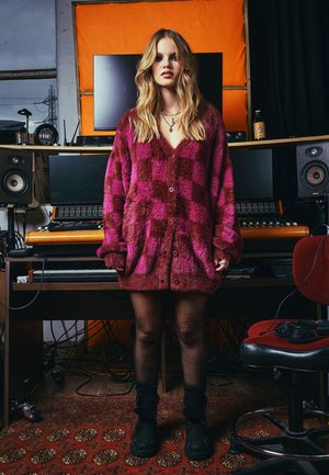Flauschige rosa und burgunderfarbene karierte Strickjacke, mit langen Ärmeln und Knöpfen vorne. Schwarze Stiefel bis zur Knöchel und dicke Socken, stehend in einem Studio-Setting.