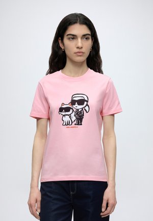 Femeie cu păr lung și închis la culoare, purtând un tricou roz cu un desen animat cu un bărbat și o pisică, amândoi purtând ochelari de soare.
