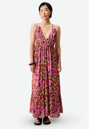 Femme debout portant une robe longue sans manches à décolleté en V profond, imprimé floral large rose, rouge et marron, et des sandales noires.