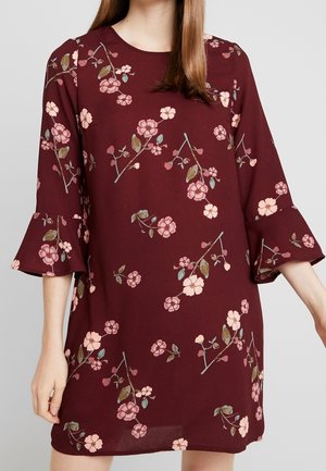 Frau trägt ein burgunderrotes Kleid mit pinken Blumenmustern und dreiviertelärmeligen Rüschenärmeln vor einem schlichten hellen Hintergrund.