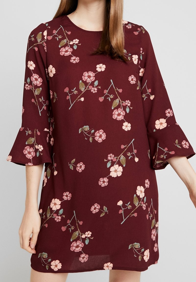 Femme portant une robe bordeaux avec des motifs floraux roses et des manches trois-quarts à volants sur un fond clair uni.
