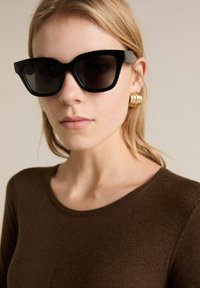 Lunettes de soleil noires surdimensionnées avec une forme géométrique élégante et des verres teintés foncés en dégradé. Le mannequin porte un haut en tricot marron et des boucles d'oreilles en or.