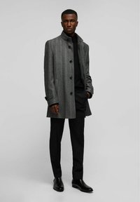 Manteau en chevrons gris avec un col mandarin, des boutons noirs et des poches latérales, associé à un pantalon noir et des bottines.